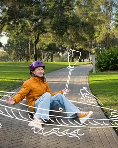 Ein Kind in einer orangen Jacke und Jeans sitzt auf einem gepflasterten Weg im Park und trägt einen lilafarbenen Helm. Um das Kind herum sind weiße Zeichnungen eines Kajaks und eines Paddels, die den Eindruck erwecken, als würde es auf Wasser paddeln.